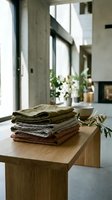 Stacked linen fabrics in neutral tones displayed on wooden table in bright modern interior with concrete walls and potted flowers for home decor styling.