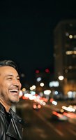 Happy adult male with dark hair and facial hair wearing black leather jacket, smiling outdoors at night with blurred city lights and urban street in background.