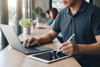 Young Asian businessman working on laptop and tablet at modern office desk with natural lighting and colleagues in background.