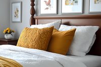 Cozy bedroom interior with dark wood headboard, white bedding, and mustard yellow accent pillows featuring textured quilting pattern and botanical wall art.