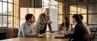 Middle-aged Caucasian male manager leading team meeting with diverse colleagues at wooden table in modern loft office with industrial windows and brick walls.