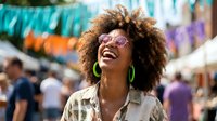 Happy young African American woman with curly afro hair and sunglasses laughing joyfully at outdoor festival with colorful decorations and crowd in background.
