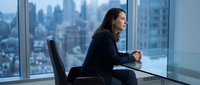 Professional businesswoman in dark suit sitting by office window overlooking city skyline, contemplating future business opportunities and career growth.