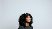 Young woman with curly black hair and medium skin tone wearing black hoodie, smiling while looking upward against white background, isolated for design use.