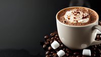 Creamy hot chocolate topped with whipped cream and cocoa powder in white ceramic cup, surrounded by coffee beans and marshmallows on dark background.