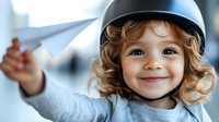 Cheerful young child wearing protective helmet smiles while playing, showing active lifestyle and safety consciousness in bright indoor setting. Curly hair peeks beneath helmet.