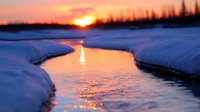 Meandering winter stream reflects golden sunset light through snowy banks, creating magical atmosphere with orange and purple hues in peaceful nature landscape.