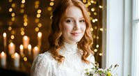 Young Caucasian bride with curly red hair in white lace wedding dress holding bouquet against golden bokeh lights background for romantic ceremony celebration.