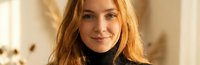 Young blonde woman with long wavy hair wearing black turtleneck sweater smiling warmly at camera in cozy indoor setting with soft natural lighting.