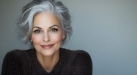 Elegant mature woman with silver gray hair wearing brown sweater smiling confidently against neutral background, representing natural aging and beauty in fashion and lifestyle.