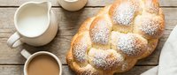 Freshly baked braided challah bread dusted with powdered sugar served with coffee and milk on rustic wooden table for breakfast or brunch meal.
