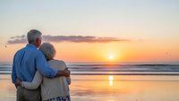 Senior couple embracing on beach at sunset watching golden hour over ocean waves. Romantic retirement lifestyle and love concept for travel destinations.