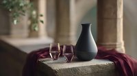 Elegant black ceramic vase and wine glasses on stone surface with burgundy draping fabric, creating moody atmospheric composition against blurred architectural background.