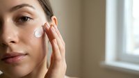 Young woman applying white facial cream moisturizer to cheek with fingers in bright bathroom for daily skincare routine and anti-aging treatment.