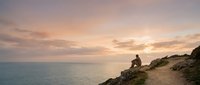 Young man sitting on rocky coastal cliff overlooking calm ocean at golden hour sunset, contemplative mood for travel, lifestyle, and wellness content.
