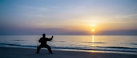 Man practicing tai chi martial arts on ocean beach at sunrise with golden sky reflection on water creating peaceful meditation atmosphere.
