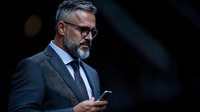Mature Caucasian businessman with gray beard and glasses checking smartphone in dark setting. Professional executive in suit displaying confidence and modern connectivity.