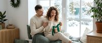 Young couple exchanging Christmas gifts in cozy living room with holiday decorations and natural light from large windows.