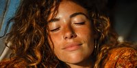 Young woman with curly hair and freckles enjoying sunlight with closed eyes, wearing orange textured clothing, conveying serenity and natural beauty.