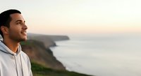 Young man with dark hair and olive complexion wearing light hoodie gazing at coastal cliffs during golden hour sunset. Serene landscape photography for travel, wellness, and lifestyle design projects.