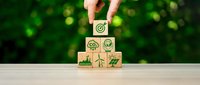 Hand placing wooden cube with green checkmark completing stack of eco-friendly icons including renewable energy, recycling symbols on natural background.