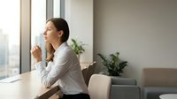 Young professional woman drinking coffee while looking out office window in modern workplace setting with natural light and plants.