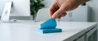 Close-up of hand placing blue sticky note on white desk surface with office supplies and plant in background, representing organization and workplace productivity.