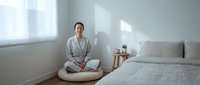 Young Asian woman in white robe practicing meditation in minimalist bedroom with natural light streaming through window creating peaceful morning atmosphere.