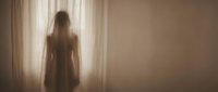 Woman with long dark hair, appearing to be of mixed ethnicity, standing in silhouette facing sheer curtains with bright light streaming through. Suitable for wellness, mental health, spirituality, and editorial design projects.