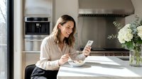 Young woman with dark hair and olive complexion, wearing a beige blouse, sitting at a modern kitchen counter holding a smartphone and smiling while enjoying breakfast. Suitable for lifestyle, technology, and wellness content.