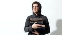 Young male student with dark hair and glasses wearing a dark hoodie, holding a stack of books against his chest, isolated on white background for educational and academic concepts.