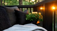 Cozy outdoor patio setting with soft cushions, knitted throw blanket, glowing lanterns and potted flowers on wooden deck railing at dusk creates relaxing ambiance.