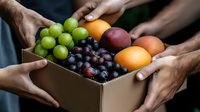 Fresh seasonal fruits in cardboard box with green and purple grapes, oranges and mango being passed between diverse hands, representing sharing and community.