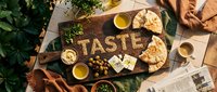 Rustic wooden board with the word Taste spelled in spices, surrounded by olives, cheese, pita bread, hummus, and fresh herbs for food blog.