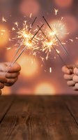 Two hands holding lit sparklers with glowing golden sparks bursting against a warm blurred background, celebrating festive occasions and special moments.