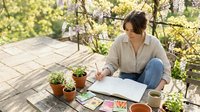 Young woman gardener planning spring garden layout while sitting on wooden deck surrounded by potted plants and seedlings in sunny backyard.
