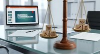 Wooden scales of justice with legal symbols on modern office desk with laptop displaying business law content for attorney consultation services.