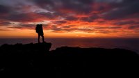 Silhouette of hiker standing on rocky mountain peak at dramatic sunset with vibrant orange and red sky, adventure and outdoor exploration concept.