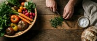 Fresh organic vegetables in wicker basket with hands chopping herbs on rustic wooden table. Healthy cooking preparation with tomatoes, carrots and greens.