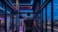 Young man with athletic build, viewed from behind, wearing black t-shirt in modern gym with floor-to-ceiling windows overlooking illuminated city skyline at dusk, neon purple and blue lighting, contemporary fitness facility interior.