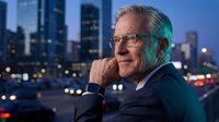 Mature male professional wearing glasses and dark business suit gazing thoughtfully at city skyline during twilight hours, suitable for corporate leadership, business success, and executive portraits.