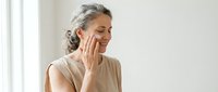 Mature woman applying skincare cream to face in bright bathroom. Anti-aging beauty routine for healthy glowing skin. Senior wellness and self-care concept.