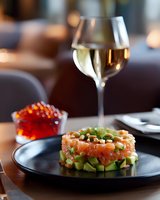 Fresh salmon tartare with diced avocado and green onions served on black plate, red caviar in crystal bowl and white wine glass in restaurant setting.