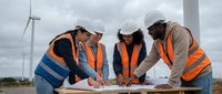 Diverse engineering team in safety gear reviewing blueprints at wind farm construction site with turbines in background for renewable energy project.