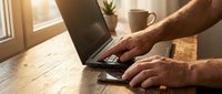 Male hands typing on laptop computer at wooden desk with coffee cup and plant in warm sunlight streaming through window creating productive home office workspace.
