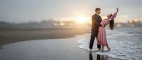 Romantic couple dancing on wet beach sand during golden hour sunset with ocean waves and soft lighting creating intimate moment for wedding or engagement.