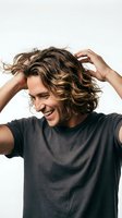 Young man with shoulder-length wavy brown hair and light complexion wearing dark casual t-shirt, running hands through hair with joyful expression against white background.