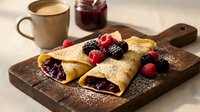 Delicious crepes filled with berry jam, topped with fresh raspberries and blackberries on wooden board. Morning breakfast with coffee and homemade preserves.