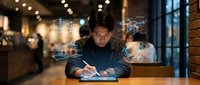 Young Asian businessman working late in modern office with holographic data displays and digital interfaces floating around him as he writes notes.