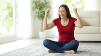Excited Asian woman celebrating success sitting cross-legged on floor at home raising fists in triumph with joyful expression in bright living room.